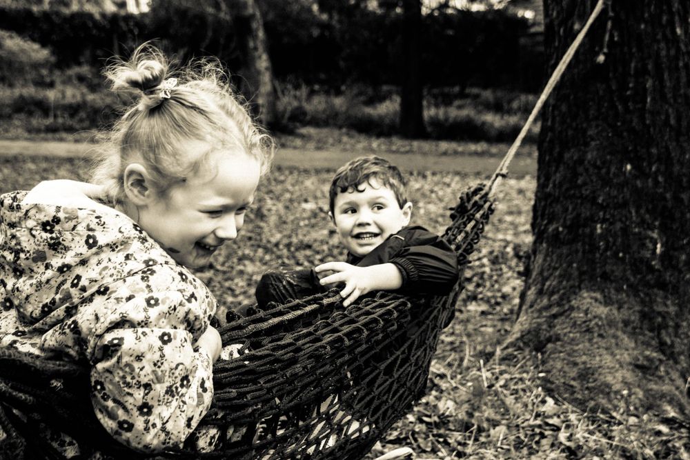 Two children play together in a dark hammock strung between trees.