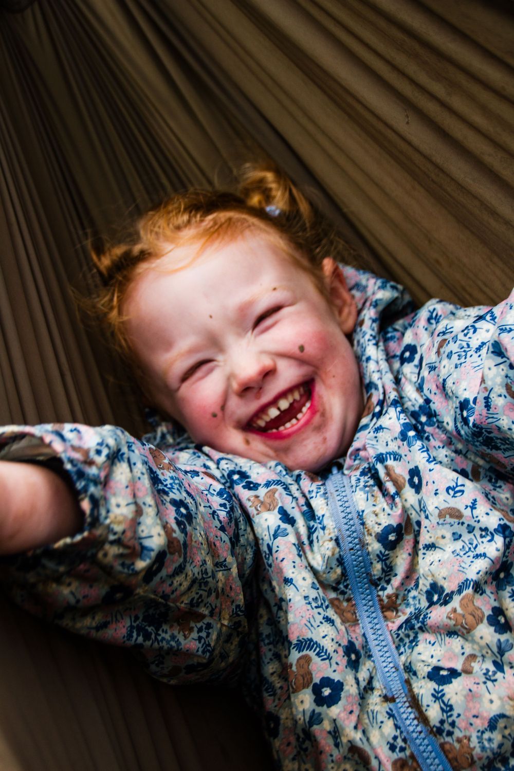 A child with red hair laughs while lying in a gray hammock outdoors.