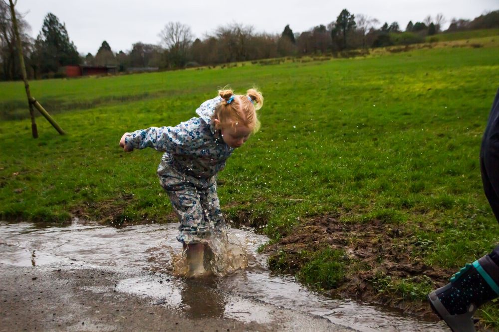 Child in rain gear jumping into puddle on muddy ground.