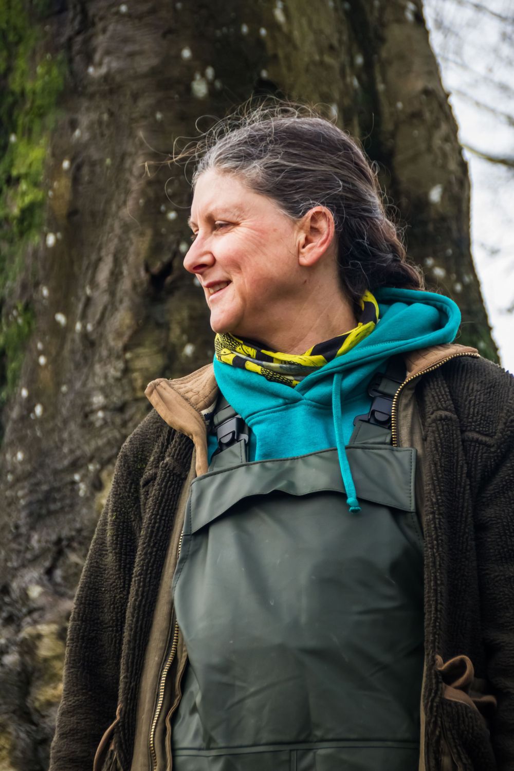 Person in outdoor gear and waders standing against mossy tree trunk.