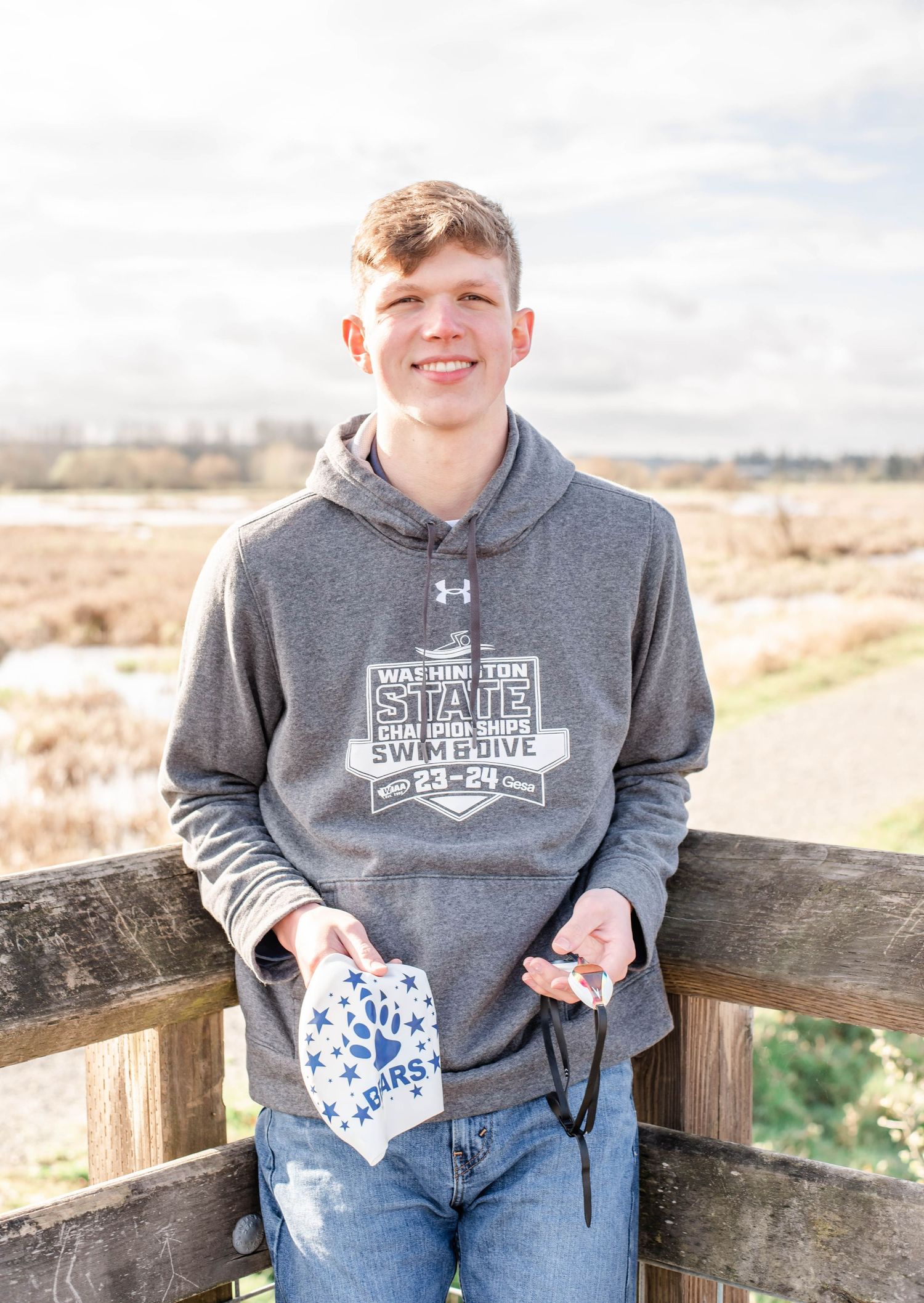 Person in grey branded hoodie standing at wooden railing with scenic background.