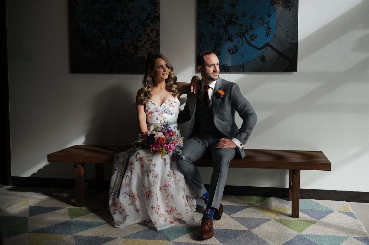 A couple sits together on a wooden bench in a modern interior with geometric flooring and artistic wall decor.