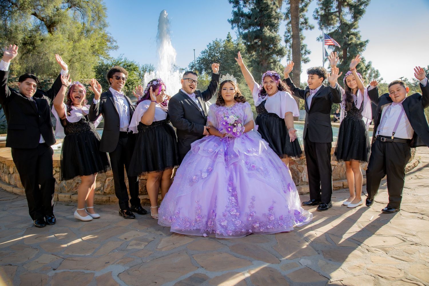 Quinceañera in purple ballgown celebrates with cheering court near fountain outdoors.