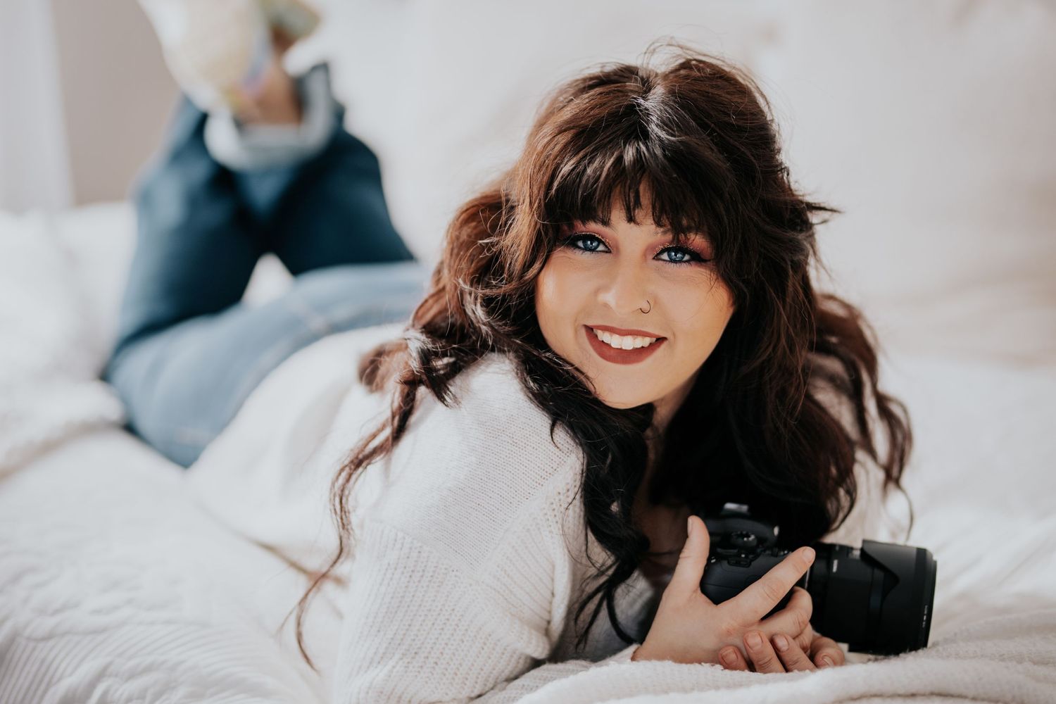 Smiling young woman with dark wavy hair lying on a white bed holding a camera in a cozy setting.