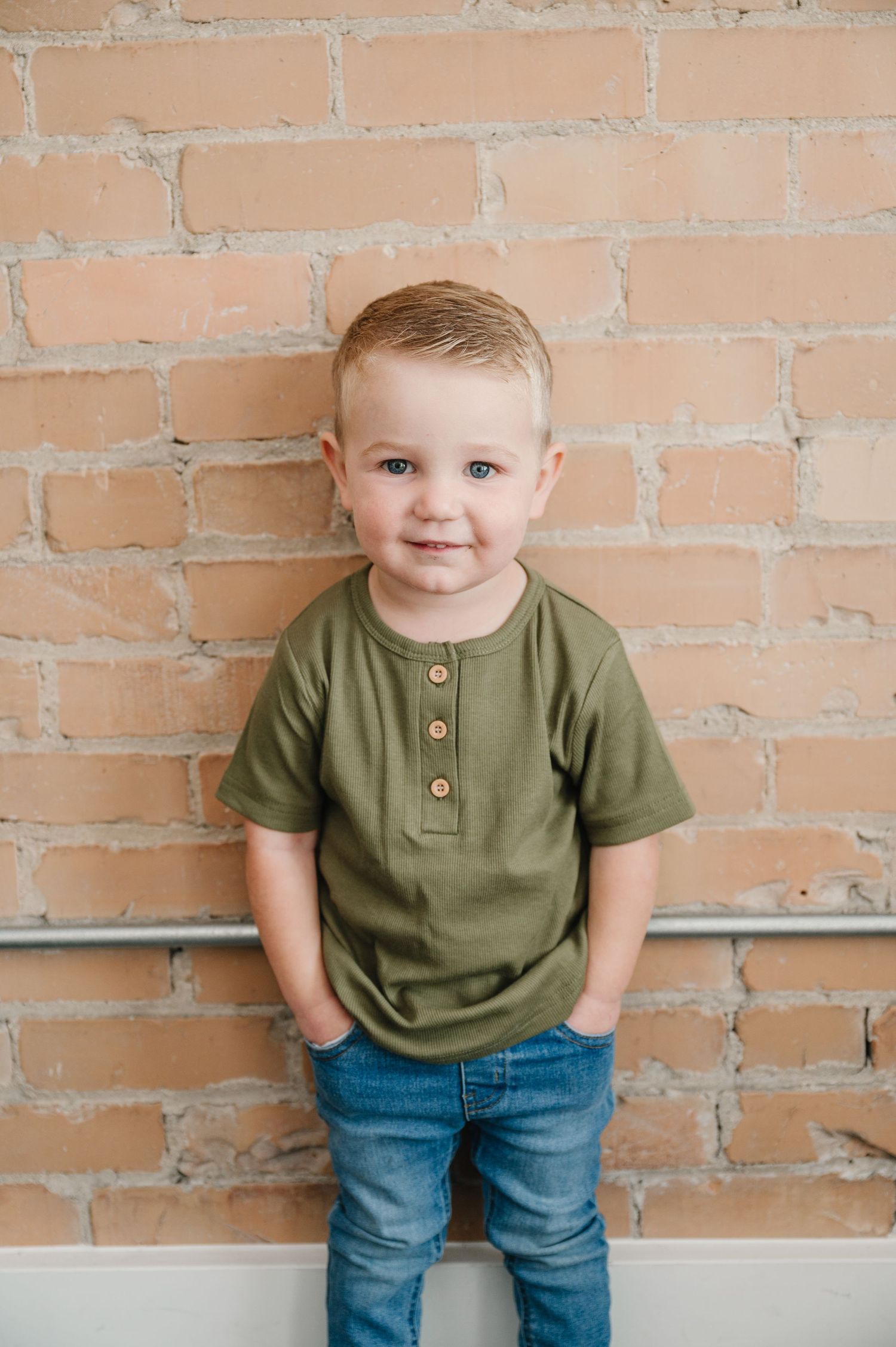 A young child wears an olive green henley shirt and blue jeans while standing against an exposed brick wall.