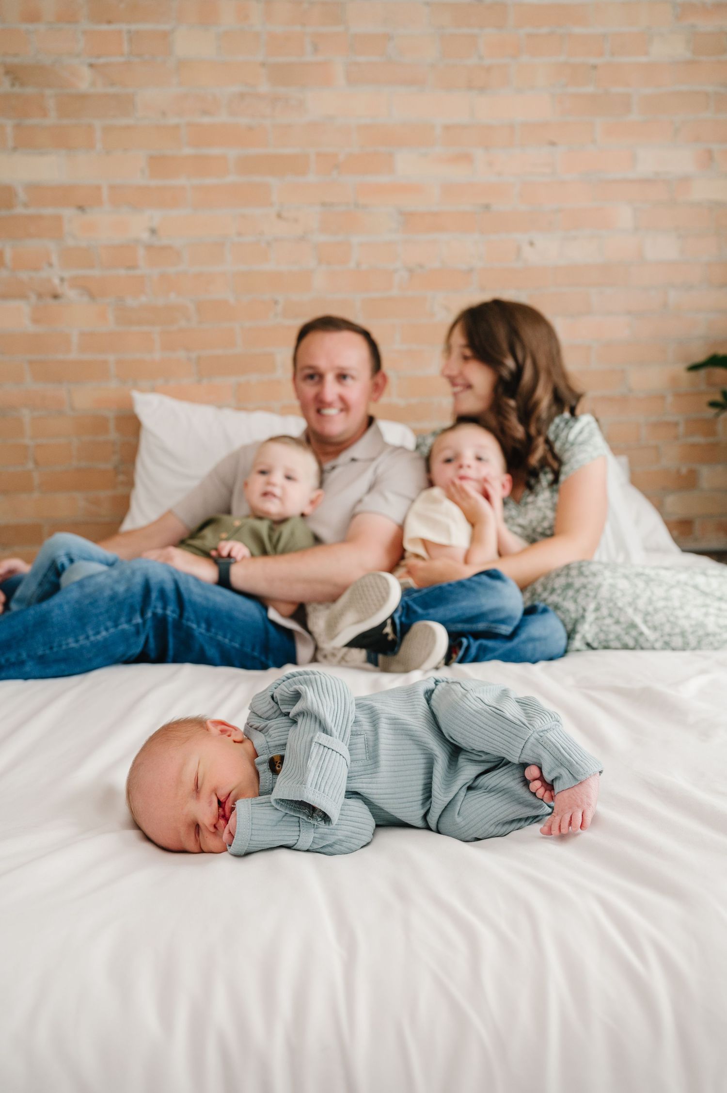 A family relaxes together on a white bed against a brick wall while a baby sleeps peacefully in the foreground.