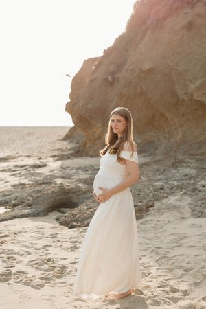 Pregnant woman in white off-shoulder gown standing on sandy beach near rocky cliffs during golden hour.