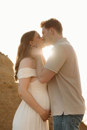 Pregnant couple sharing a kiss at golden hour, woman in white off-shoulder dress, man in beige shirt and jeans.