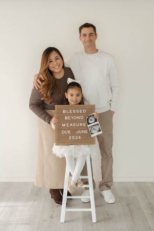 Pregnancy announcement photos showing wooden sign saying 'Beyond Measure' with ultrasound and white stool against neutral wall.