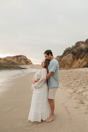 A couple in white and blue embraces on a scenic beach with rocky cliffs during golden hour.