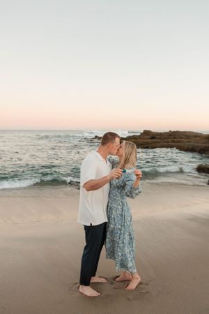 A couple in casual attire dances playfully together on a sandy beach at sunset with ocean waves in the background.