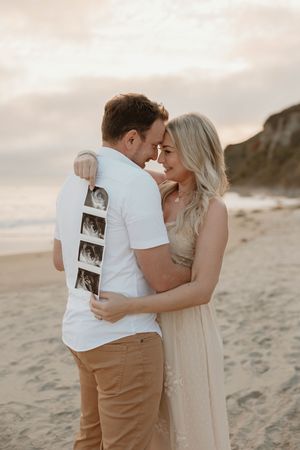 A couple embraces tenderly on a sandy beach at sunset holding an ultrasound photo.