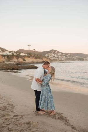 A couple shares a romantic moment at sunset on a scenic beach with mountains in the background.