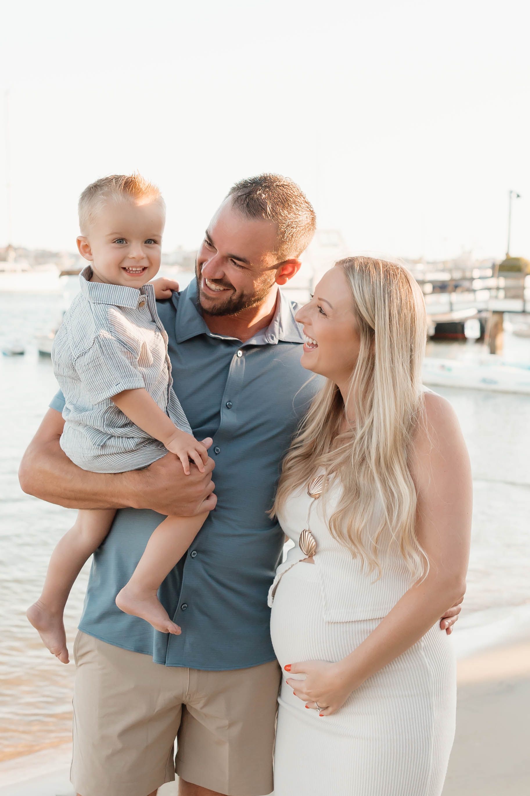 A happy family poses by the marina, with a man holding a toddler boy while a pregnant blonde woman smiles beside them.