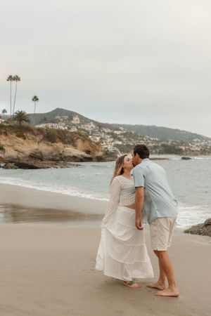 A couple shares a romantic moment on a scenic beach with coastal cliffs and mountains in the background.
