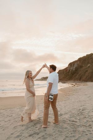 Couple celebrating joyfully during sunset beach photoshoot in soft neutral tones.