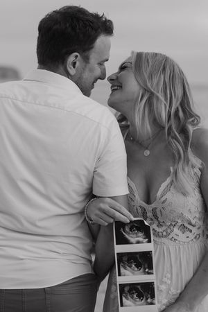 A couple in white outfits share a romantic moment while holding ultrasound photos in black and white.