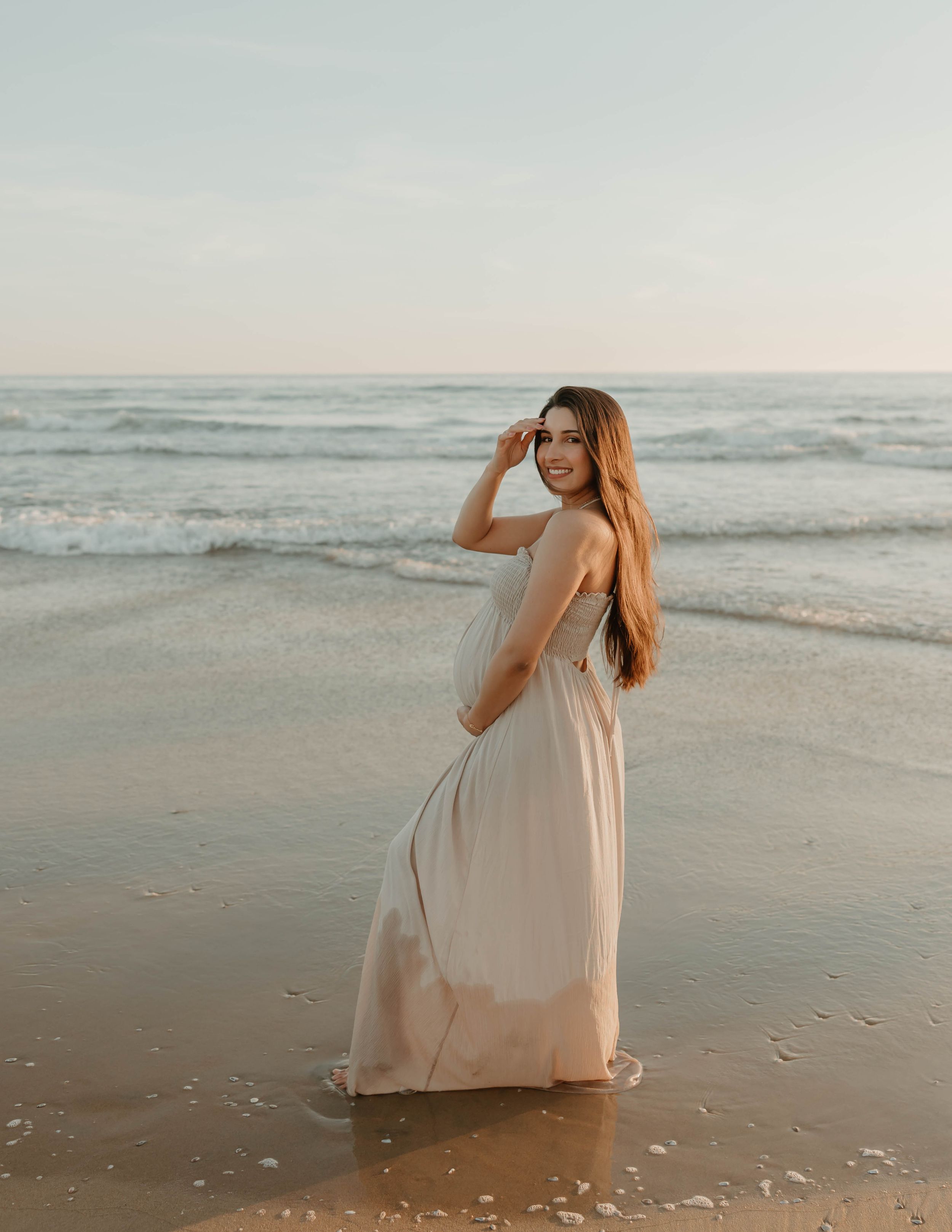 A person in a flowing beige dress poses gracefully on a serene beach at sunset with waves in the background.