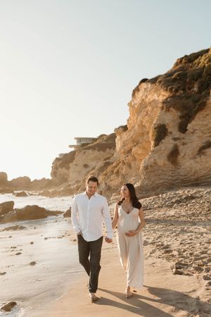 Couple walking together along a rocky beach with cliffs at sunset creating a romantic coastal atmosphere.