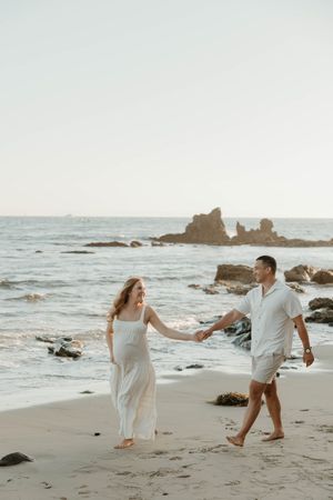 A couple walks hand in hand along a rocky beach at sunset with waves crashing against coastal rock formations.