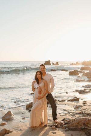 A couple shares a romantic moment on a rocky beach at sunset as waves crash against the shore.