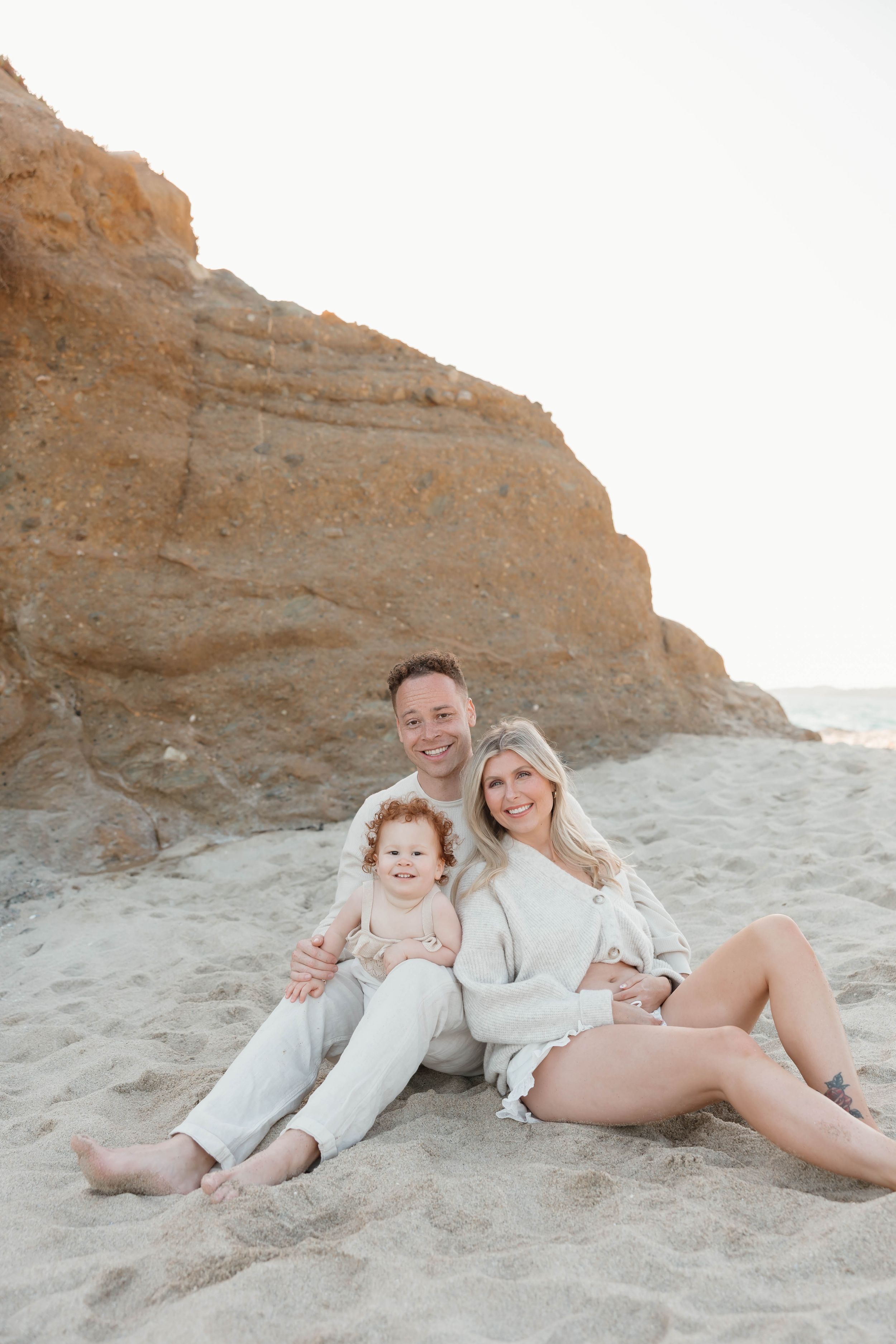 A happy family of three sits on sandy beach near rocky cliffs, parents smiling with their young toddler in soft neutral tones.