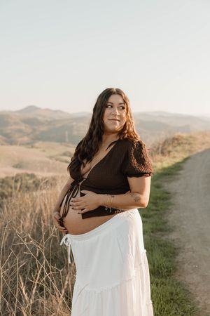 Expectant mother in black top and white skirt stands on hilltop trail during golden hour sunset in rolling hills.