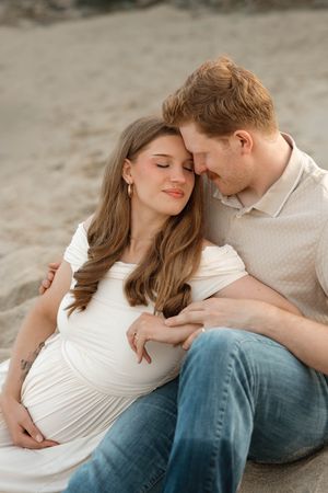 A pregnant woman in a white dress sits with her partner on sandy ground, both smiling with foreheads touching tenderly.