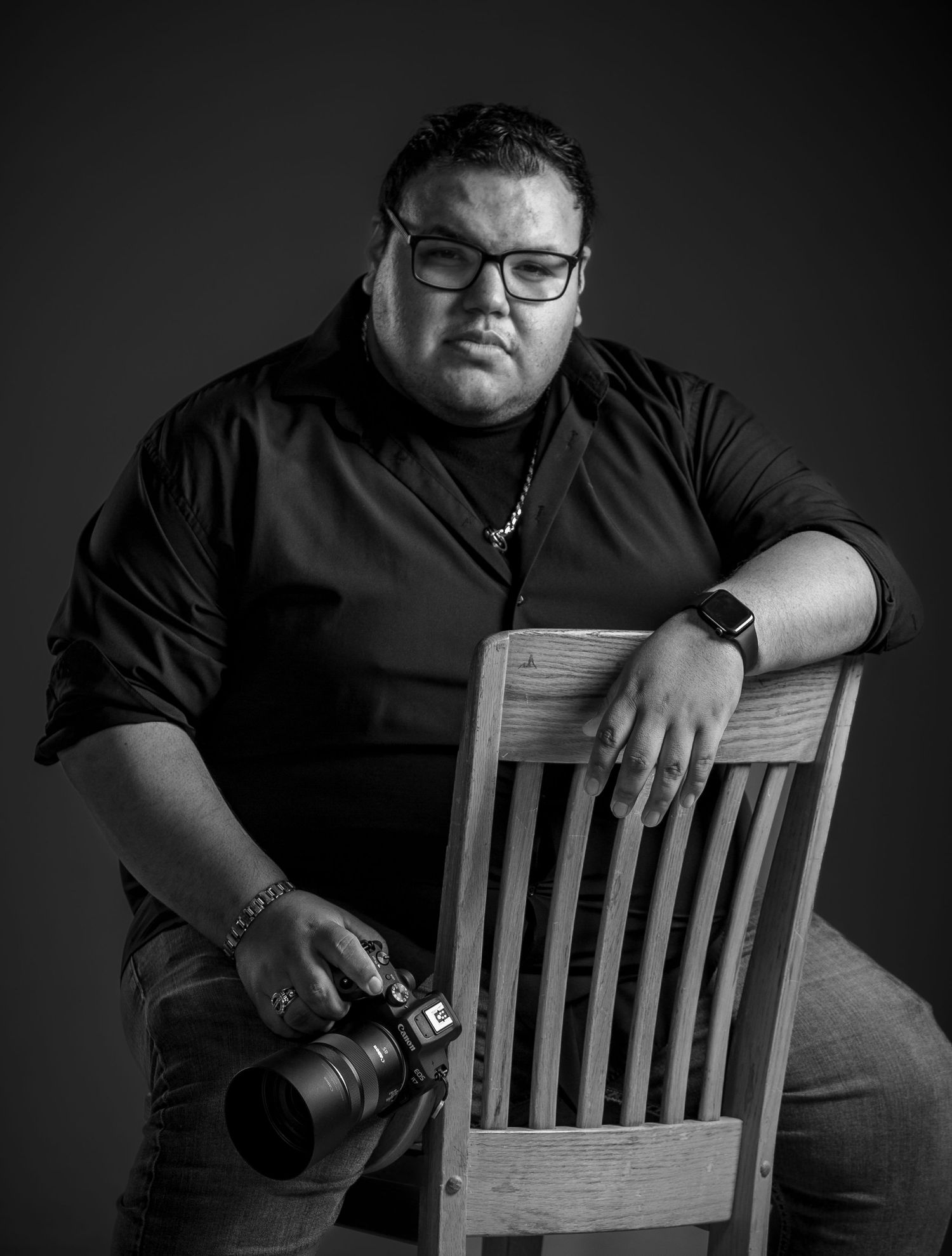 Black and white portrait of photographer sitting on wooden chair holding camera equipment.