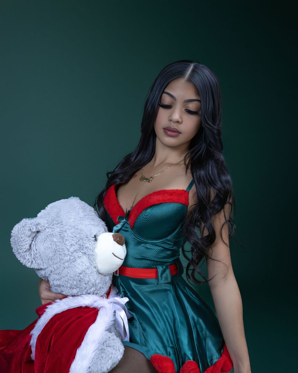 Model in emerald green Christmas dress with red trim poses with white teddy bear wearing Santa outfit against dark background.
