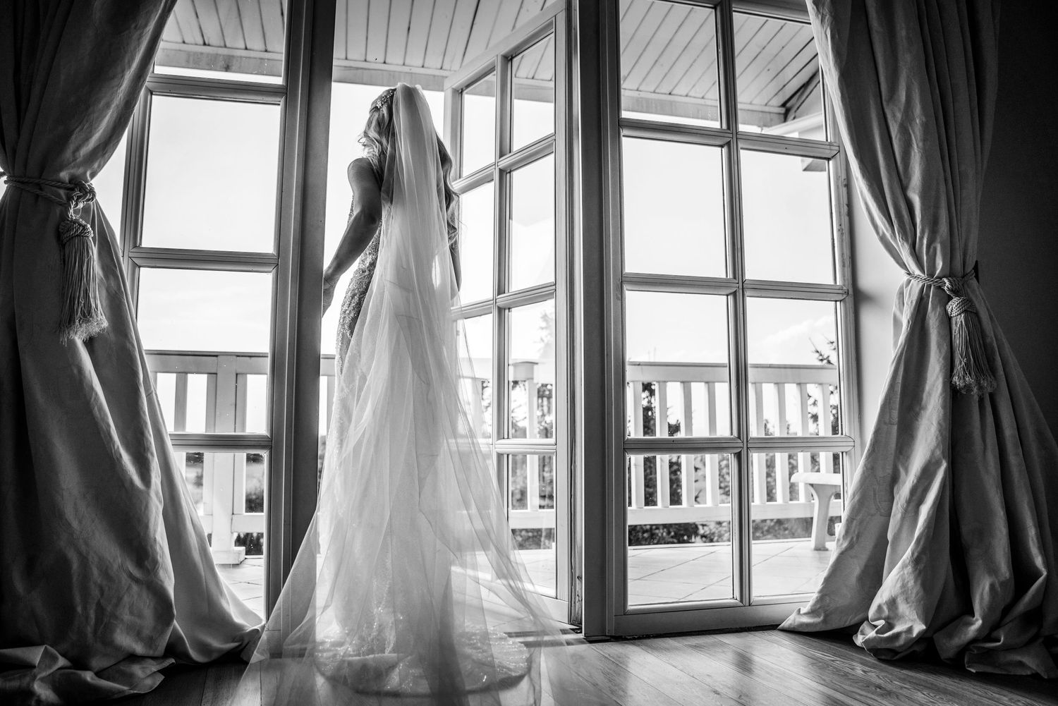 A silhouetted figure in a wedding dress stands by large windows with flowing curtains.