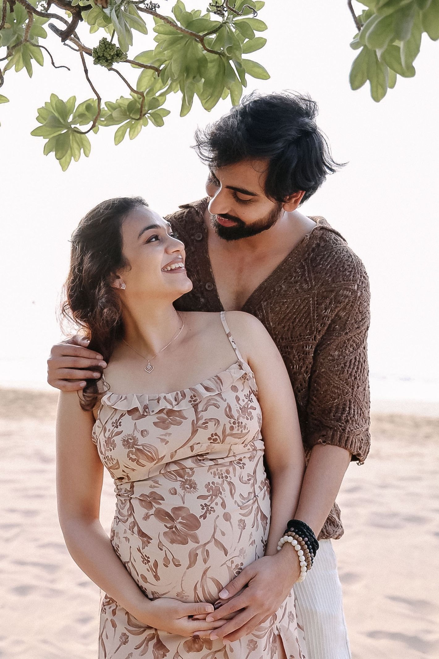 A couple embraces outdoors under green foliage, with her wearing a floral dress and him in a brown sweater.
