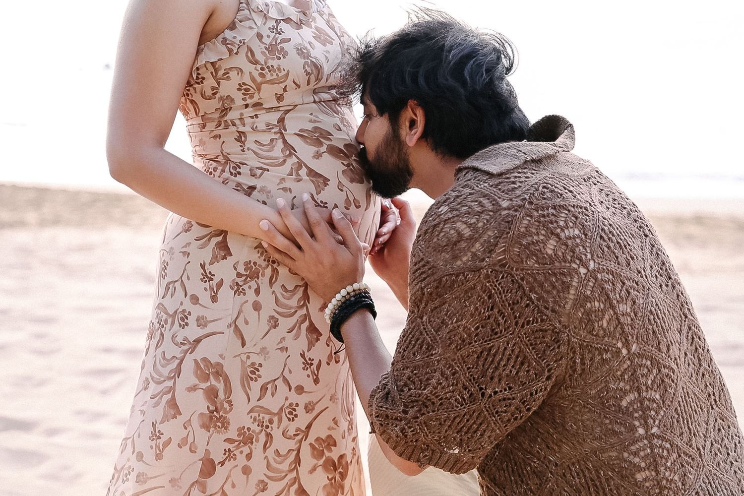 Romantic couple embraces on sandy beach at sunset wearing floral dress and brown sweater.