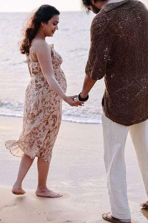 A couple stands together on a beach, with the woman wearing a flowy floral dress and the man in white pants.