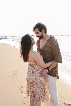 Intimate beach photoshoot of couple in floral dress and brown sweater against ocean backdrop.