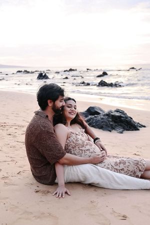 A couple in romantic poses on a sandy beach with rocky coastline during sunset.