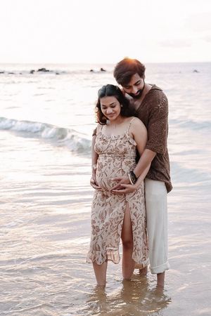 A couple stands together in shallow ocean water during sunset wearing beige and brown outfits.