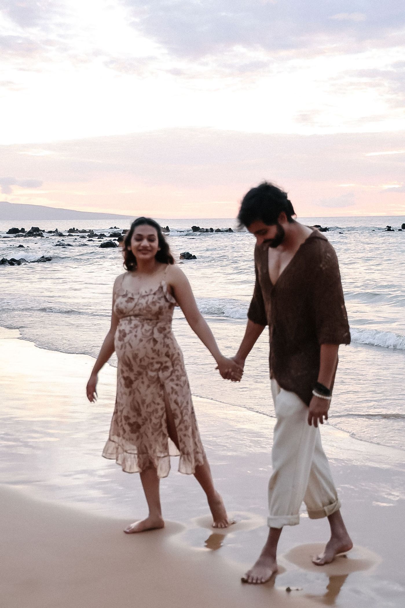 A couple takes a romantic stroll along the shoreline at dusk with the ocean and rocks in the background.