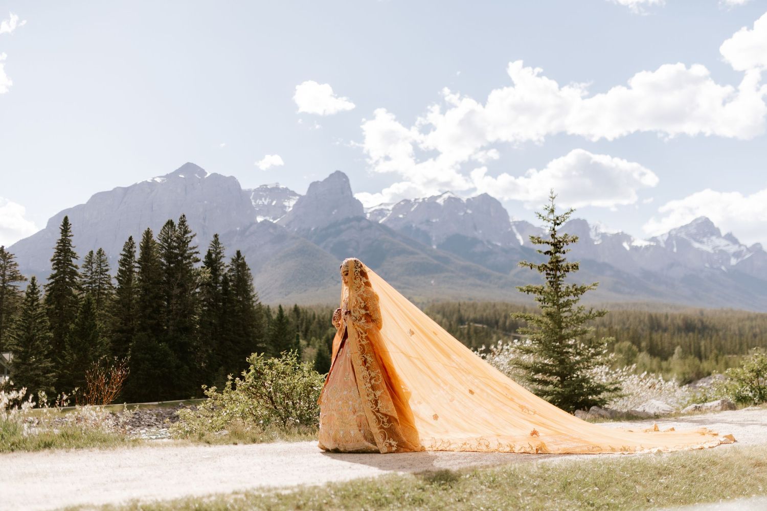 Orange tent pitched in mountainous landscape with evergreen trees and snow-capped peaks in background.