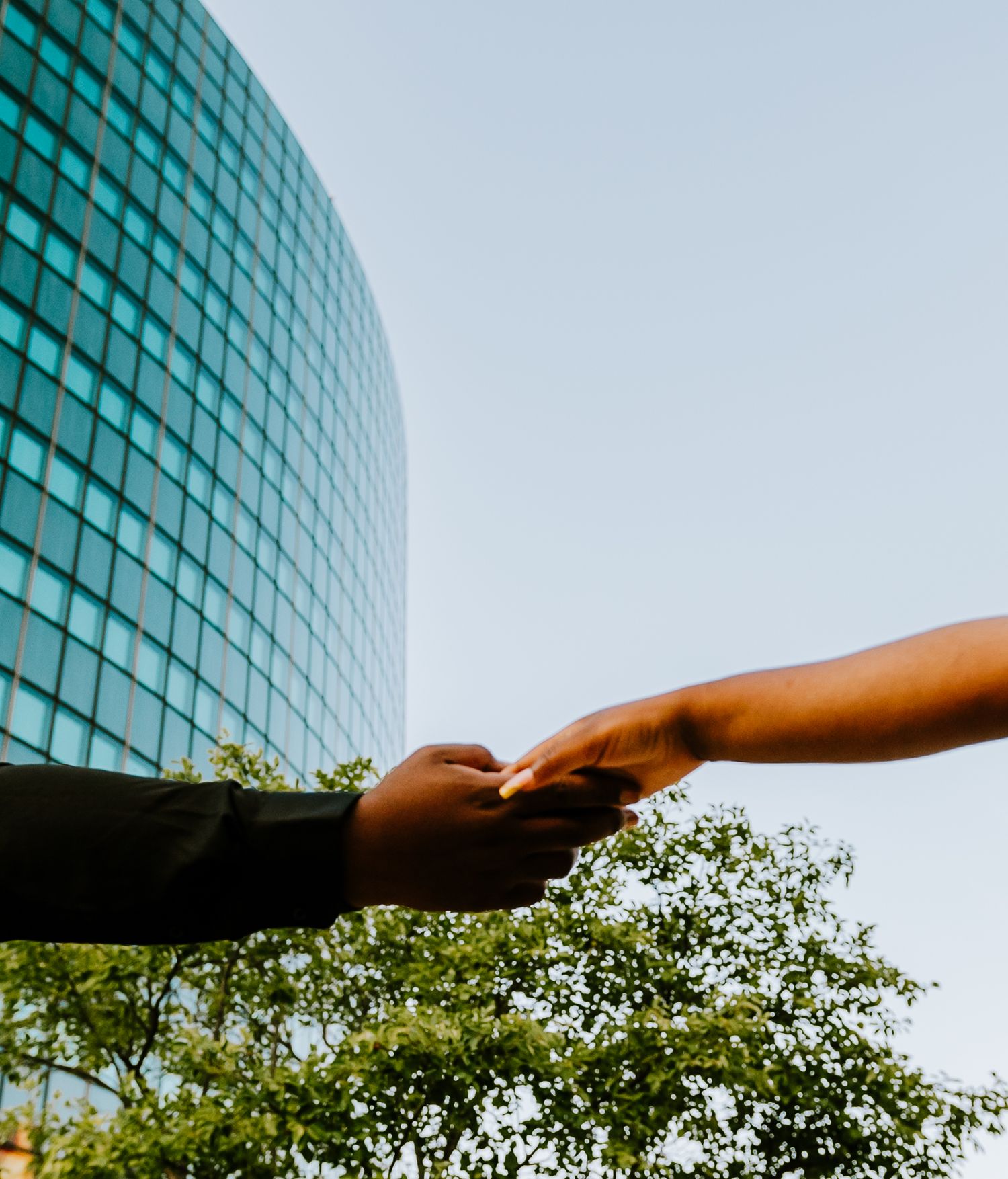 Two hands reach towards each other in front of a modern glass building and trees against a blue sky in Hartford, Connecticut.