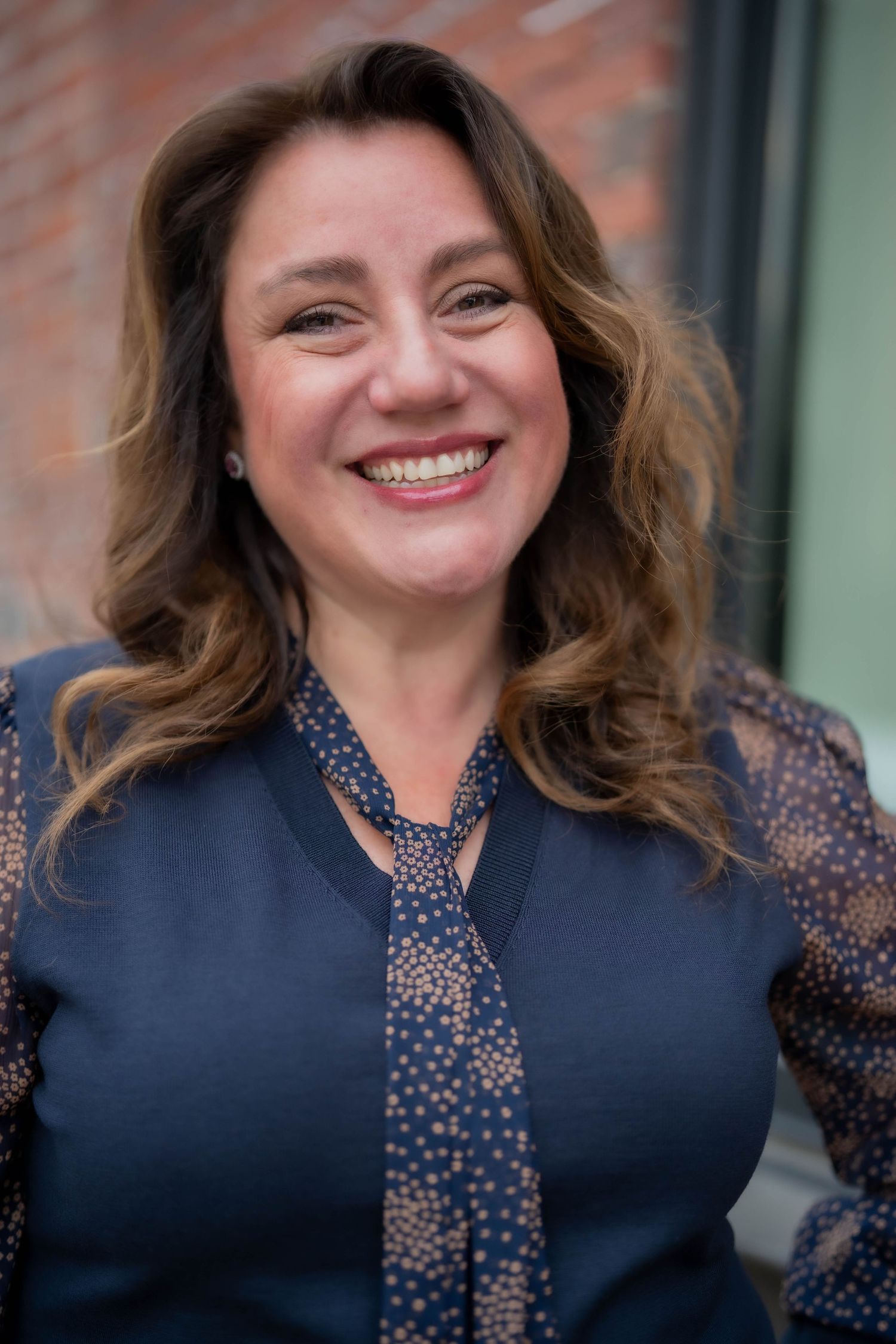 Smiling woman with brown wavy hair wearing a navy blazer and patterned blouse, standing outdoors near a brick wall.