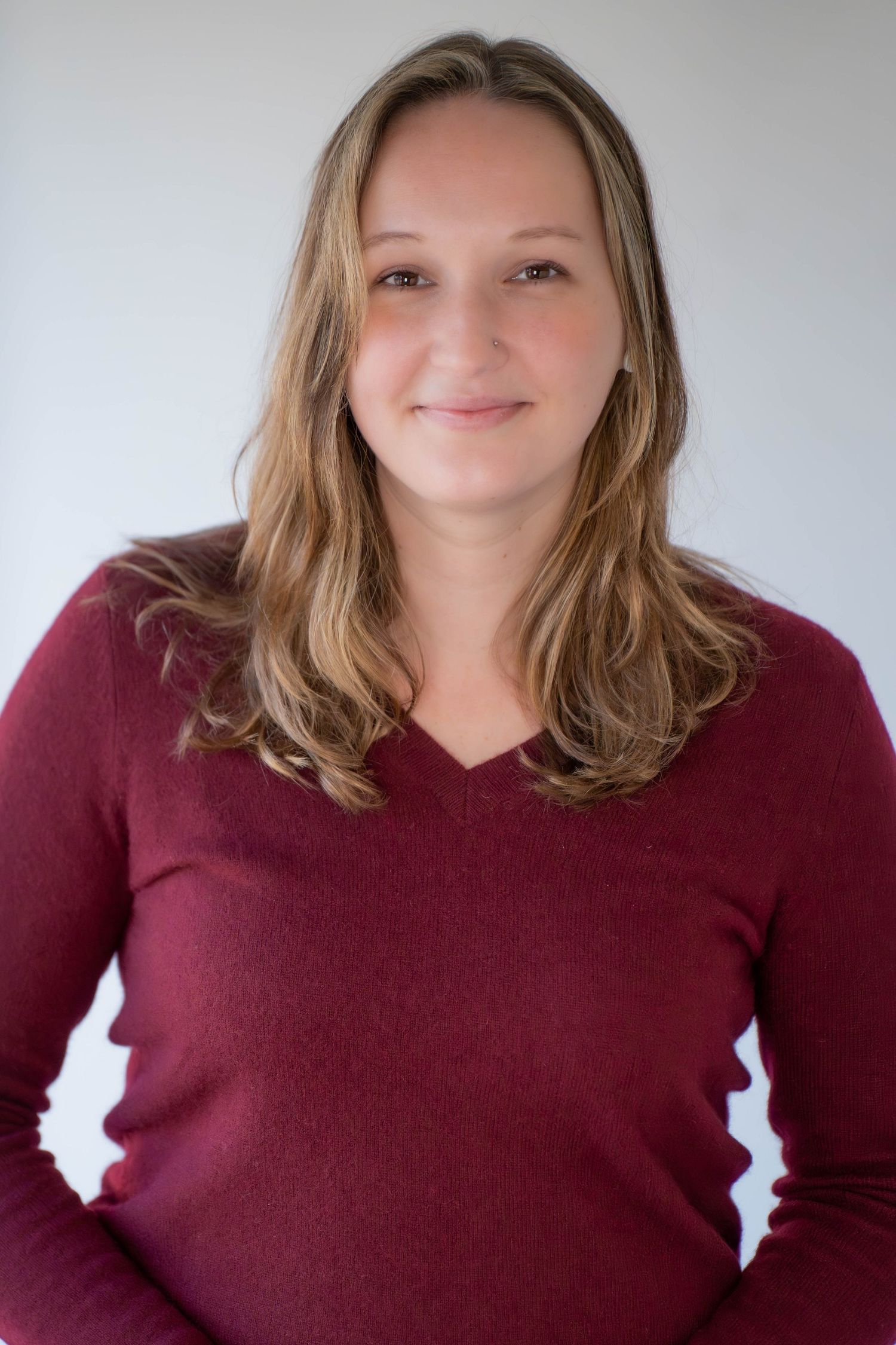 Woman in burgundy v-neck sweater smiling warmly against a light gray background.