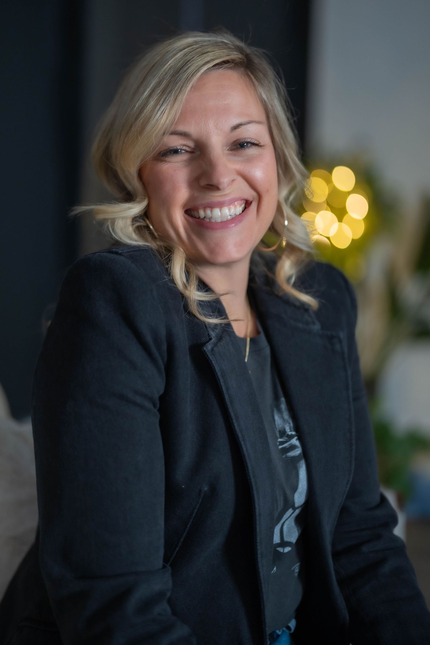 Smiling blonde woman in a dark blazer, posing confidently with bokeh lights in the background.