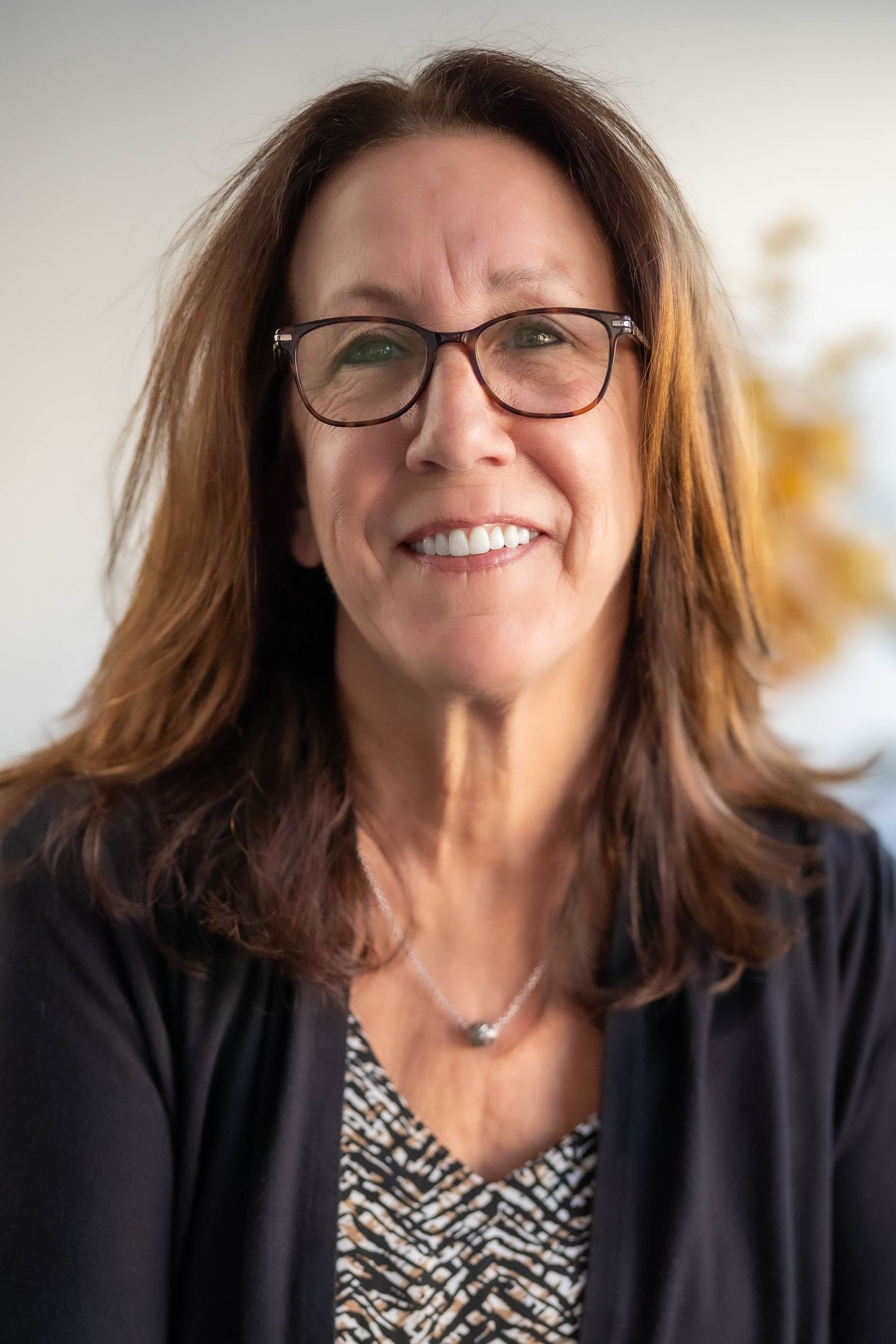 Smiling middle-aged woman with brown hair and glasses wearing a dark blazer and patterned top in a professional headshot.