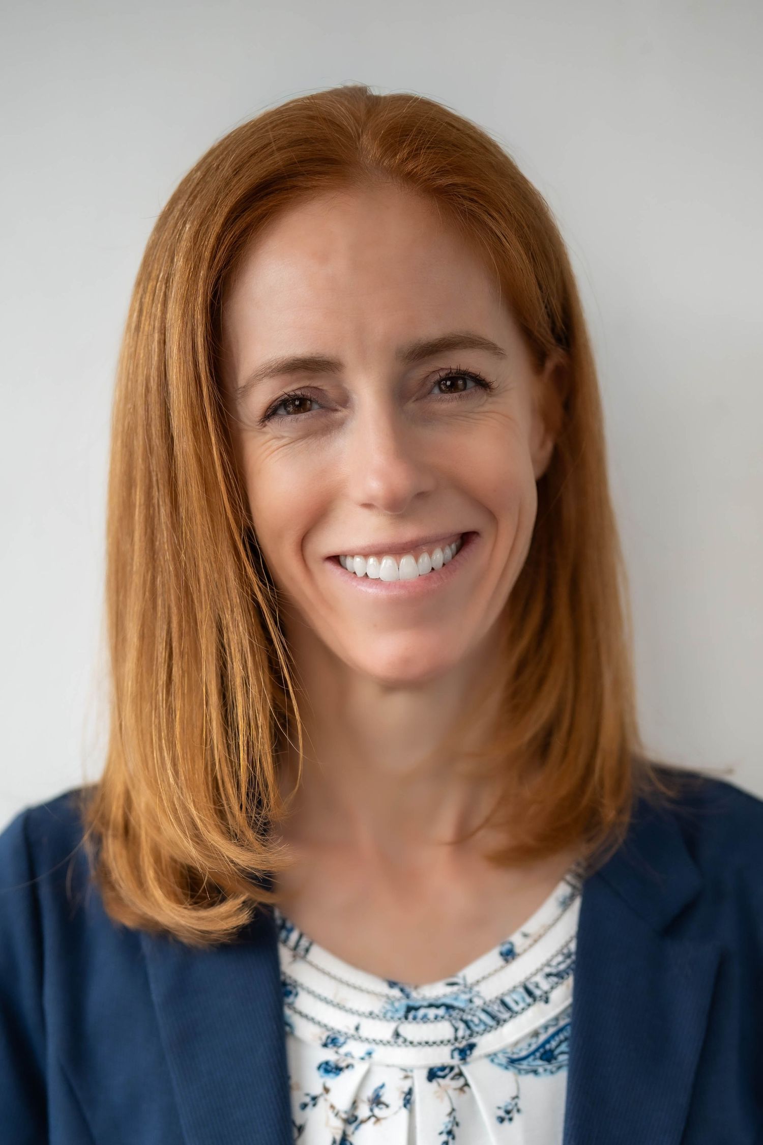 Professional headshot of a smiling woman with red hair wearing a blue blazer and floral blouse.