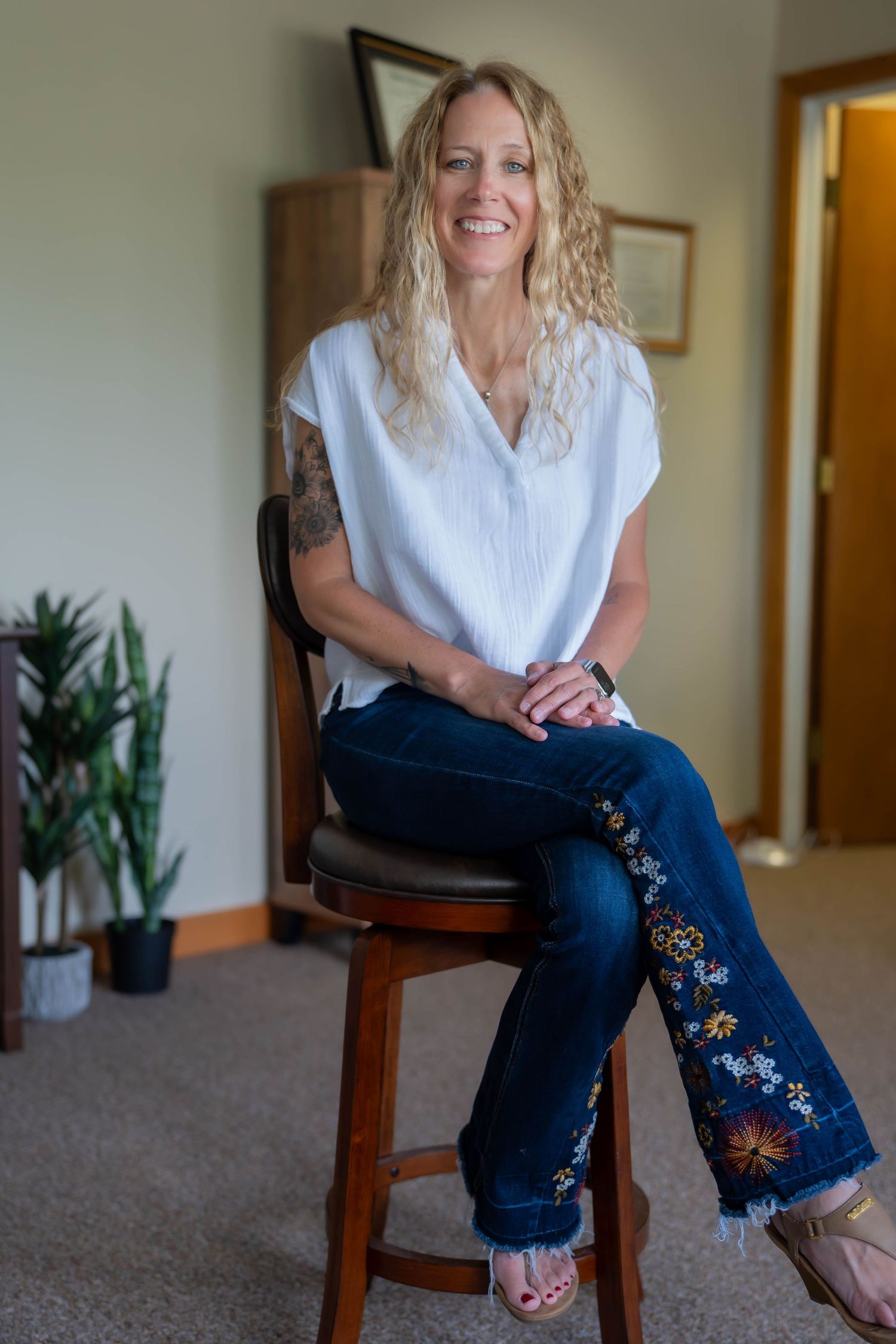 Smiling blonde woman with tattoo sitting on wooden stool wearing white top and floral embroidered jeans indoors.
