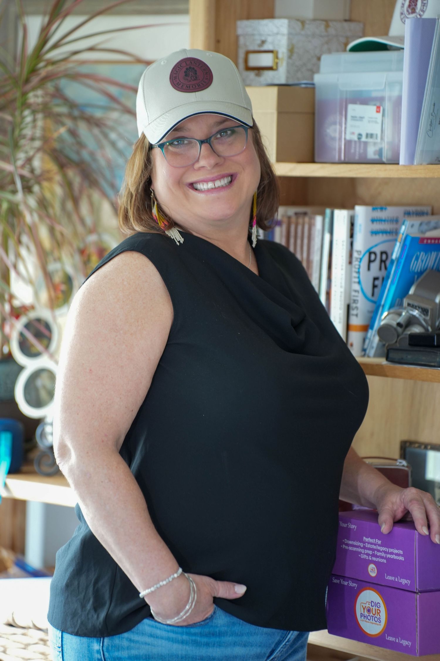 Smiling woman in black sleeveless top, glasses, and white cap poses by wooden shelves in a cozy shop.