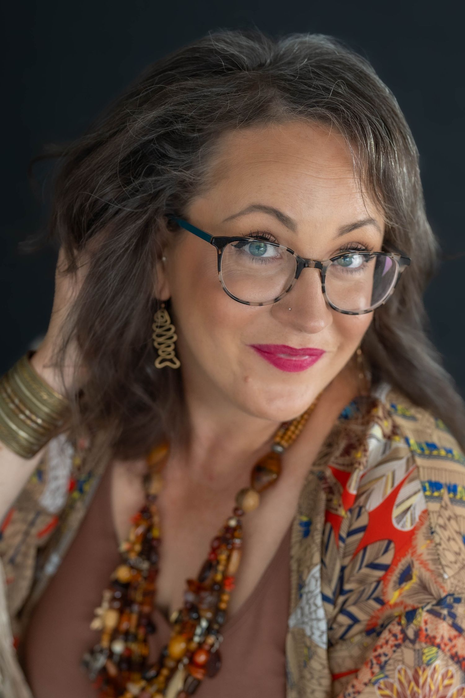 A stylish middle-aged woman with blue eyes, glasses, red lips, layered necklaces, and a colorful patterned jacket poses confidently.