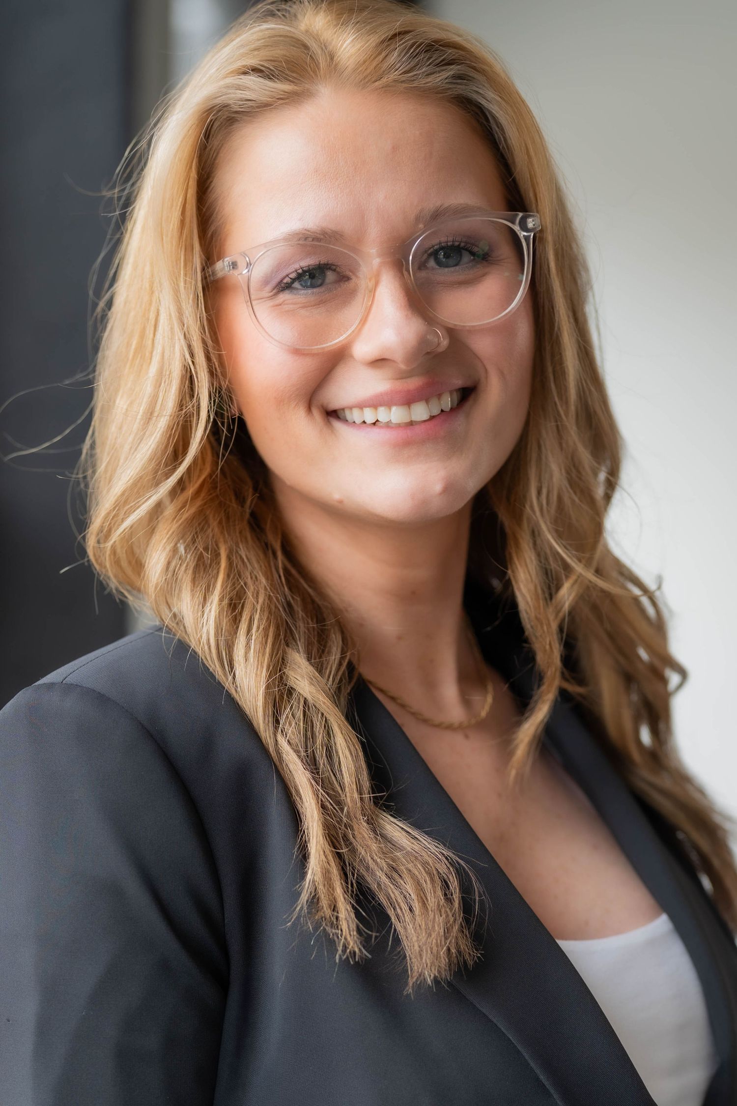 Smiling young professional woman with blonde hair and clear glasses wearing a gray blazer in a modern office setting.