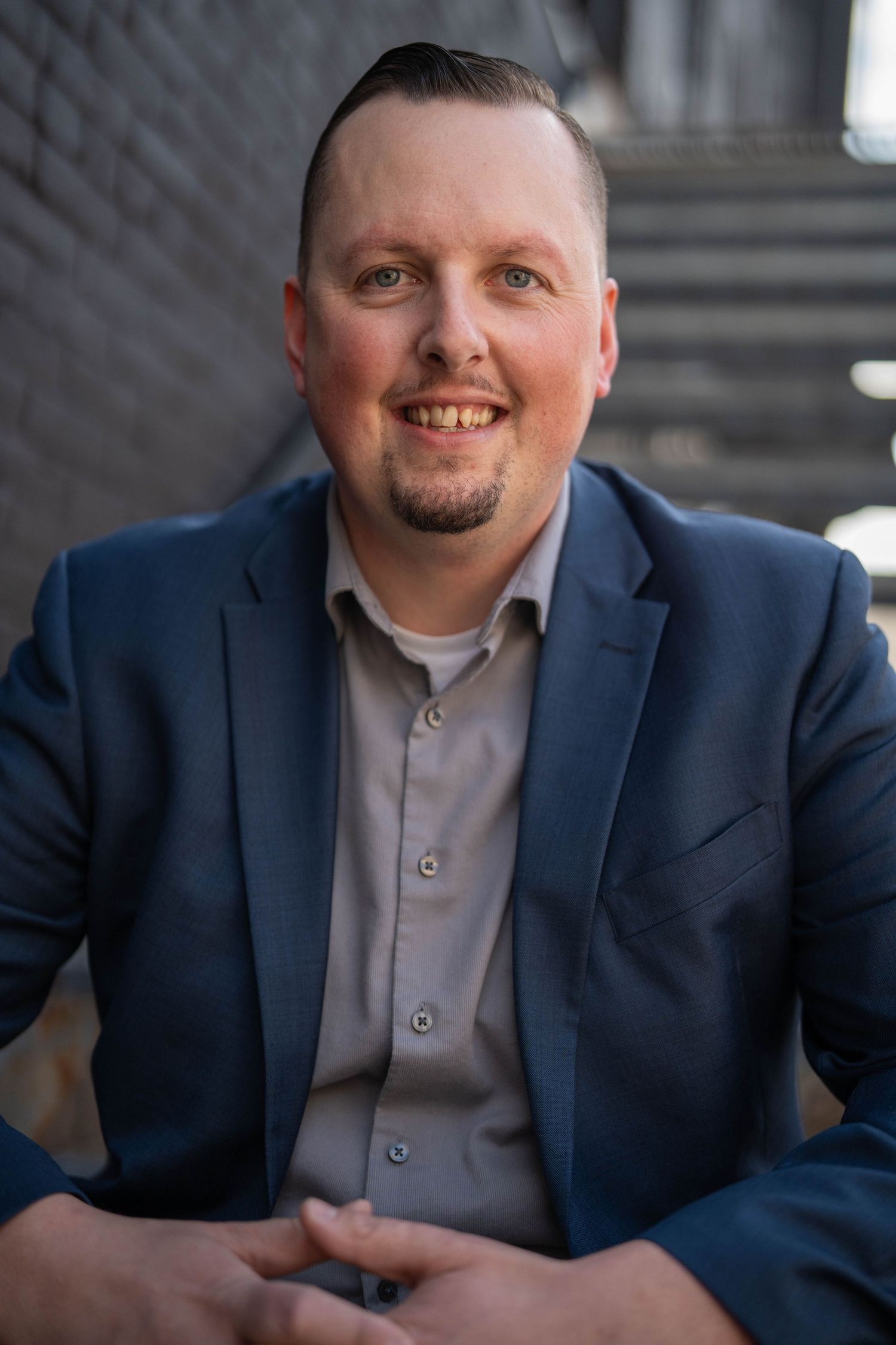 Smiling man in blue blazer and gray shirt seated outdoors, with a well-groomed beard and friendly expression.